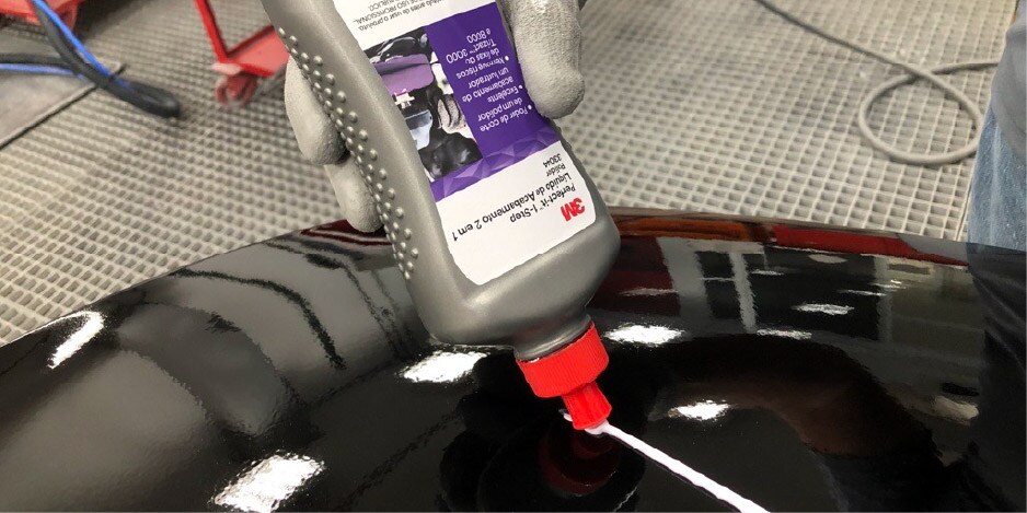 Pessoa aplicando composto de polimento 3M em uma superfície automotiva preta e brilhante dentro de uma área de oficina.

