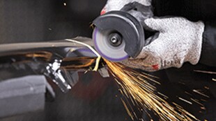 Uma mão com luva operando uma ferramenta rotativa para desbaste em metal, gerando faíscas durante o processo
