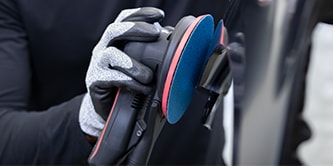 Trabalhador usando luvas de proteção segurando uma lixadeira com um disco abrasivo azul enquanto se prepara para lixar a superfície de um veículo.
