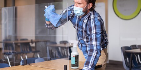 Imagem de um homem com camisa listrada e máscara limpando um separador de plástico em uma sala de aula.
