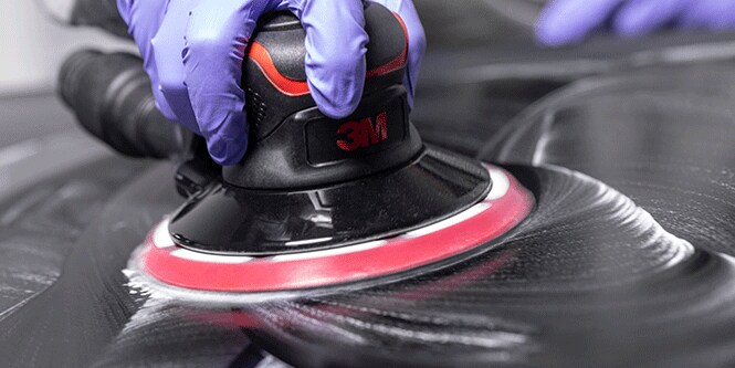 Uma pessoa usando luvas roxas utiliza uma lixadeira elétrica da marca 3M em uma superfície escura, criando um padrão em espiral.
