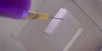 Video still of a needle inserting dyed liquid between bonded plastic panels to show resistance to moisture intrusion
