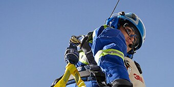 Worker hanging in sky in safety harness wearing clear, fog-free protective eyewear, hardhat, safety vest and hearing protection
