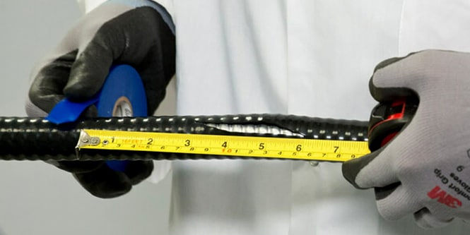 Pessoa usando luvas de proteção medindo uma haste metálica com superfície rugosa usando uma fita métrica e segurando uma fita de marcação azul.
