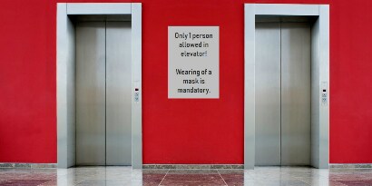 Dois elevadores com uma placa entre eles, com o seguinte escrito (traduzido do inglês): “Apenas 1 pessoa permitida no elevador! O uso de máscara é obrigatório.”
